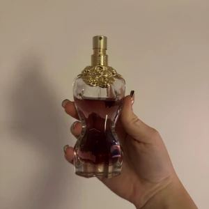 Parfymflaska med gulddekor - Säljer en elegant parfymflaska med en unik form som liknar en kvinnokropp. Flaskan är genomskinlig med en vacker röd vätska inuti och har en lyxig guldfärgad dekor på toppen. Perfekt för den som vill ha något både stilfullt och unikt på sminkbordet.