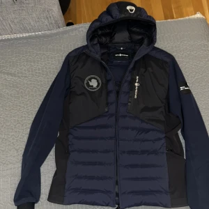 Mörkblå jacka från Sail Racing - Snygg mörkblå jacka från Sail Racing med huva och dragkedja. Jackan har ett stilrent märke på bröstet och är perfekt för kyligare väder. Den är långärmad och har en quiltad design för extra värme, perfekt för vinter. Skick 9/10 priset kan diskuteras storlek S, men passar även Xs och M