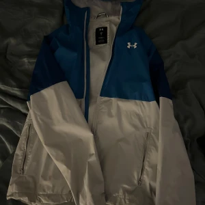 Under Armour Jacka - Snygg blå och vit jacka från Under Armour med huva och dragkedja. Perfekt för utomhusaktiviteter med sin sportiga design och funktionella fickor. Enda defekten är fläckarna som syns på bilden, går säkert bort med en bra tvätt, men har aldrig testat