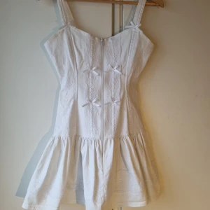 Vit klänning med rosetter - Charmig vit klänning med axelband och söta rosetter framtill. Klänningen har en volangdetalj vid fållen och ett diskret mönster i tyget. Perfekt för somriga dagar.