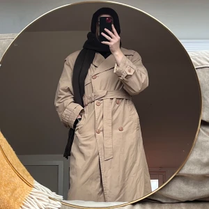 Beige trenchcoat med bälte - Beige trenchcoat med klassisk design och bälte i midjan. Jackan har knappar framtill och långa ärmar. Perfekt för höst och vår.
