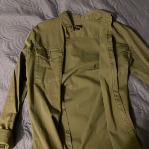 Olivgrön skjorta från Zara Man - Snygg olivgrön skjorta från Zara Man med långa ärmar och knappar framtill. Skjortan har två bröstfickor med knappdetaljer och en klassisk krage. Perfekt för en stilren look.