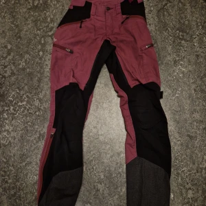 Vinröda byxor från Lundhags - Säljer ett par vinröda byxor/vandrarbyxor från Lundhags i storlek 36. De har svarta förstärkningar på knän och insida ben, samt praktiska dragkedjor på fickorna. Perfekta för utomhusaktiviteter och äventyr!