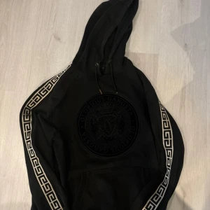 Svart hoodie från Glorious Gangsta - Säljer en svart hoodie från Glorious Gangsta i storlek M. Den har ett coolt mönster längs ärmarna och en stor ficka framtill. Perfekt för en avslappnad stil eller streetwear-look. Huvan är justerbar med snören. Använd ett par gånger men i bra skick.