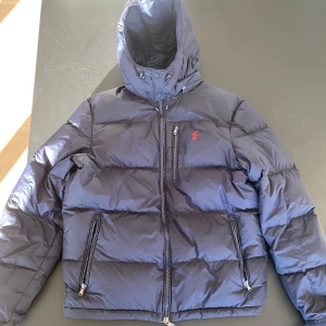 Mörkblå dunjacka från Polo Ralph Lauren - Säljer en snygg mörkblå dunjacka från Polo Ralph Lauren. Jackan har en dragkedja framtill och två praktiska fickor med dragkedjor. Inte andvänd mycket och med ingen odör.