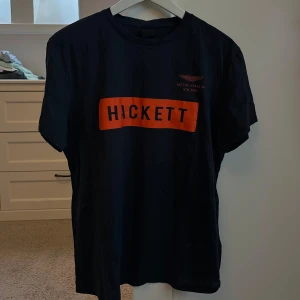 Svart t-shirt från Hackett - Säljer en svart t-shirt från Hackett med Aston Martin Racing-logga. Den har en stor orange text på framsidan. Perfekt för vardagsbruk eller för att visa ditt intresse för racing. T-shirten är i bra skick och har en normal passform.