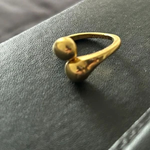Guldfärgad ring fån Pilgrim med unik design - Snygg guldfärgad ring med en modern och unik design. Ringen har två sfäriska ändar som möts på ovansidan, vilket ger den ett stilrent och elegant utseende. Perfekt för att ge en touch av glamour till din outfit.