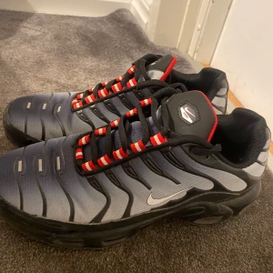 Nike Air Max Plus Tn - Snygga Nike Air Max Plus Tn sneakers i svart och grått med röda detaljer. Skorna har en unik design med synlig Air Max-dämpning och mönstrade snören. Perfekta för en sportig look.
