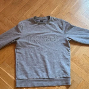 Grå sweatshirt från Hugo Boss - Säljer en stilren grå sweatshirt från Hugo Boss med broderad logga på framsidan. Tröjan har en klassisk passform och är perfekt för en avslappnad look. Tillverkad i mjukt material för extra komfort.