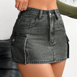 Svart jeanskjol med fickor - Trendig svart jeanskjol med stora fickor på sidorna och fram. Kjolen har en kort längd och stängs med dragkedja och knapp framtill. Perfekt för en cool och avslappnad stil.