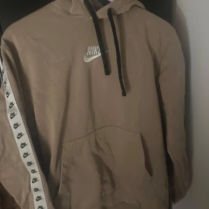 Beige hoodie från Nike - Säljer en snygg beige hoodie från Nike i bra skick. Den har en klassisk Nike-logga på bröstet och svarta snören i huvan. Ärmarna har en cool detalj med små Nike-loggor längs sidan. Perfekt för höst och vår!