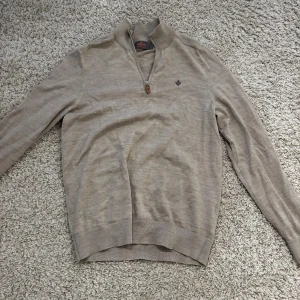 Beige tröja från Morris - Säljer en stilren beige tröja från Morris i 100% merinoull. Tröjan har en dragkedja vid halsen och ett diskret broderat emblem på bröstet. Perfekt för både höst och vår. Den är i mycket bra skick och passar till både vardag och fest.