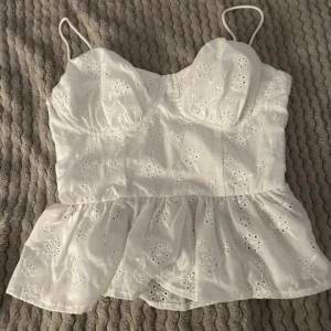 Vit ärmlös topp med spetsdetaljer - Söt vit topp med tunna axelband och spetsdetaljer. Den har en volangkant nedtill. Toppen är i mycket bra skick och passar till både jeans och kjol.