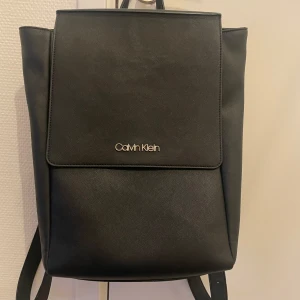 Svart ryggsäck från Calvin Klein - Snygg och stilren svart ryggsäck från Calvin Klein i mycket bra skick. Tillverkad i syntetmaterial med ett minimalistiskt design. Perfekt för både vardag och mer formella tillfällen. Ryggsäcken har justerbara axelremmar och ett stort huvudfack med plats för det mesta och även en bärbardator 