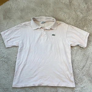 Vit pikétröja från Lacoste - Säljer en klassisk ljusrosa pikétröja från Lacoste i bra skick. Tröjan har korta ärmar och den ikoniska krokodilloggan på bröstet. Perfekt för en avslappnad men stilren look. Passar både till vardags och lite finare tillfällen. 🐊