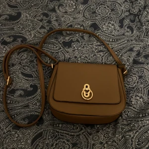 Brun axelväska med gulddetaljer - Snygg brun axelväska i skinn med gulddetaljer. Väskan har en justerbar axelrem och ett elegant spänne framtill. Insidan har flera fack med dragkedja för smidig organisering. Perfekt för både vardag och fest!