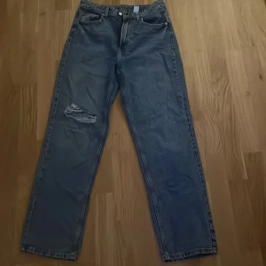 Blå jeans med slitningar - Säljer ett par klassiska blå jeans i bra skick. De har en cool slitning på ena benet och en lös passform. Perfekta för en avslappnad stil. Passar till både vardag och fest!