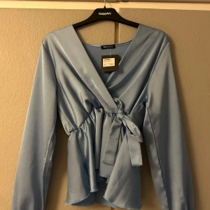 Ljusblå omlottblus från Missay - Säljer en elegant ljusblå omlottblus från Missay i sidenliknande material. Blusen har långa ärmar och ett knytband i midjan för en smickrande passform. Perfekt för både vardag och fest!