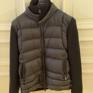 Moncler jacka/västjacka - Säljer en snygg svart pufferjacka från Moncler med stickade ärmar. Jackan har en dragkedja framtill och två praktiska fickor med dragkedjor. Perfekt för höst och vinter, håller dig varm och stilren. Moncler-loggan finns på ärmen. Passar både till vardags och lite finare tillfällen. Allt kan diskuteras ✅