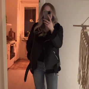 Svart pilot jacka från NA-KD - Snygg svart jacka från NA-KD med dragkedja och bälte. Jackan har en cool bikerstil med långa ärmar och är perfekt för höst och vinter. Den är tillverkad i ett material som liknar skinn och har en mjuk insida.  