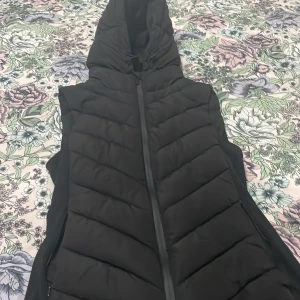 Svart dunväst från Zara - Snygg svart dunväst från Zara med dragkedja och huva. Perfekt för kyliga dagar när du vill hålla dig varm och stilren. Västen har en quiltad design och är lätt att matcha med olika outfits. Bra för vinter 