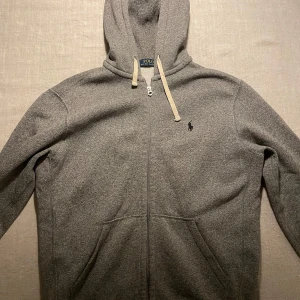 Grå hoodie från Ralph Lauren - Säljer en snygg grå hoodie från Ralph Lauren i mycket bra skick. Den har en dragkedja framtill och en liten broderad logga på bröstet. Perfekt för höst och vår, med en skön passform och huva med snören. Passar både till vardags och träning!