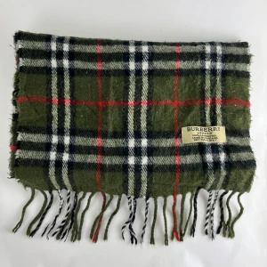 Grön rutig halsduk från Burberry - Säljer en snygg grön rutig halsduk från Burberry i 100% lammull. Den har klassiska fransar i ändarna och ett stilrent mönster i svart, vitt och rött. Perfekt för att hålla sig varm och trendig under kyliga dagar. Tillverkad i England och endast kemtvätt.