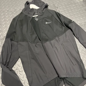 Svart löparjacka från Nike - Säljer en svart Nike löparjacka i Therma-Fit material, perfekt för kyliga dagar. Jackan har en dragkedja framtill och är långärmad med en stilren design. Den är i mycket bra skick och passar perfekt för löpning eller andra utomhusaktiviteter.
