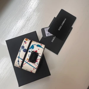 Fodral för hörlurar från Dolce & Gabbana - Snyggt fodral för hörlurar från Dolce & Gabbana för iPods promed ett färgglatt stänkmönster i blått, rött och gult på en vit bakgrund. Fodralet har en elegant design med en liten svart platta med logotypen på framsidan. Perfekt för att ge dina hörlurar en stilfull touch. (Ons i pods ingår inte;)