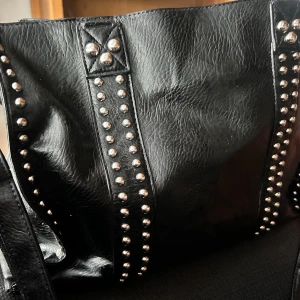 Vintageväska i skinn! - Äkta stor och rymlig Vintageväska i skinn med dekorativa nitar, justerbar axelrem och ett stort fack som stängs med knäppknapp. Mått: B: 40 cm H: 30 cm D: 15 cm. Den är i fantastiskt skick för sin ålder och jag har smort den med läderbalsam och lagt in en bottenplatta. Den är hel och ren utan skavanker.