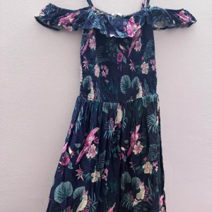 Blommig offshoulder klänning - Charmig offshoulder klänning med blommigt mönster i rosa och grönt på en mörkblå bas. Klänningen har volangdetaljer vid axlarna och smala axelband. Perfekt för sommarens varma dagar.