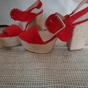 Röda platåsandaler med stabil klack - Snygga röda sandaler med hög klack och platåsula i repmaterial. Sandalerna har en bred rem över foten och en justerbar rem med spänne runt ankeln. Perfekta för att ge lite extra höjd och stil.