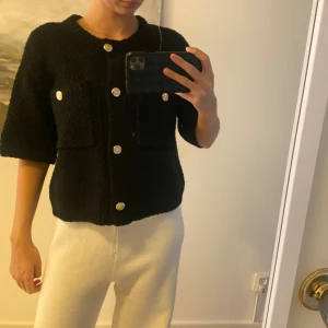 Svart topp med knappar - Snygg svart topp med trekvartsärm och stora knappar. Toppen har två framfickor och en textur som ger ett stilrent intryck. Perfekt för en trendig look.