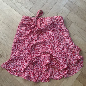 Röd blommig omlottkjol från Indiska - Säljer en söt röd omlottkjol med vita blommor från Indiska. Kjolen har volangdetaljer och en omlott-design som ger en fin passform. Perfekt för sommarens varma dagar! 🌸jag har knappt använt kjolen och därmed är den i mycket bra skick. Priset kan diskuteras🥰💕