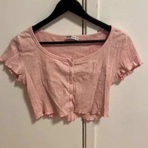 Rosa ribbad croppad topp från FB Sister - Säljer en söt rosa croppad topp från FB Sister. Toppen är ribbad med korta ärmar och knappar framtill. Perfekt för en avslappnad stil.