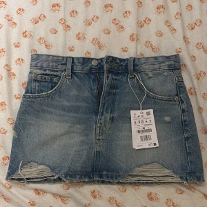 Jeanskjol från Pull&Bear - Snygg jeanskjol från Pull&Bear i klassisk blå denim. Aldrig använt den var för liten på mig. Kjolen har en rå kant nertill och slitna detaljer för en trendig look. Den har fem fickor och stängs med dragkedja och knapp framtill. Perfekt för en avslappnad stil. 