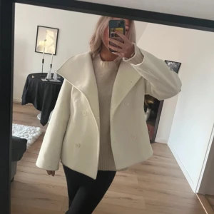 Vit kappa - Säljer en stilren vit kappa med dubbelknäppning och bred krage. Jackan har en elegant och tidlös design som passar perfekt för kyligare dagar. Den är långärmad och har en klassisk passform.