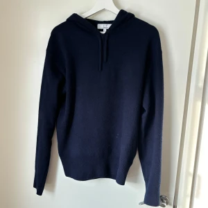 Mörkblå hoodie i kashmir från Carmés - Säljer en lyxig mörkblå hoodie från Carmés i 100% kashmir. Den har en klassisk design med huva och dragsko, perfekt för kyliga dagar. Tröjan är mjuk och bekväm med långa ärmar och ribbade muddar.