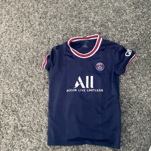 Mörkblå PSG fotbollströja - Säljer en mörkblå PSG fotbollströja med korta ärmar och vit text på framsidan. Tröjan har röda och vita detaljer runt halsen och ärmsluten. Perfekt för fotbollsfans!