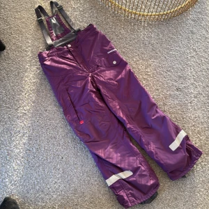Lila skidbyxor med hängslen - Säljer ett par lila skidbyxor med justerbara hängslen. Byxorna har reflexdetaljer och förstärkningar vid bensluten. Det är en liten reva på vänster sida nere vid foten på 1,5 cm och på höger sida på 0,5cm. Perfekta för vinteraktiviteter.