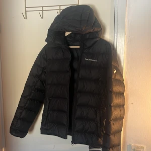 Svart dunjacka från Peak Performance - Säljer en svart dunjacka från Peak Performance. Jackan har en huva och dragkedja framtill. Perfekt för kyliga dagar med sin vadderade design som håller dig varm. En stilren och funktionell jacka för alla tillfällen.
