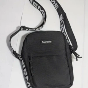 Svart axelväska från Supreme - Snygg svart axelväska från Supreme med justerbar axelrem och dragkedja. Tillverkad i slitstarkt Cordura-material. Perfekt för att bära dina dagliga nödvändigheter med stil.
