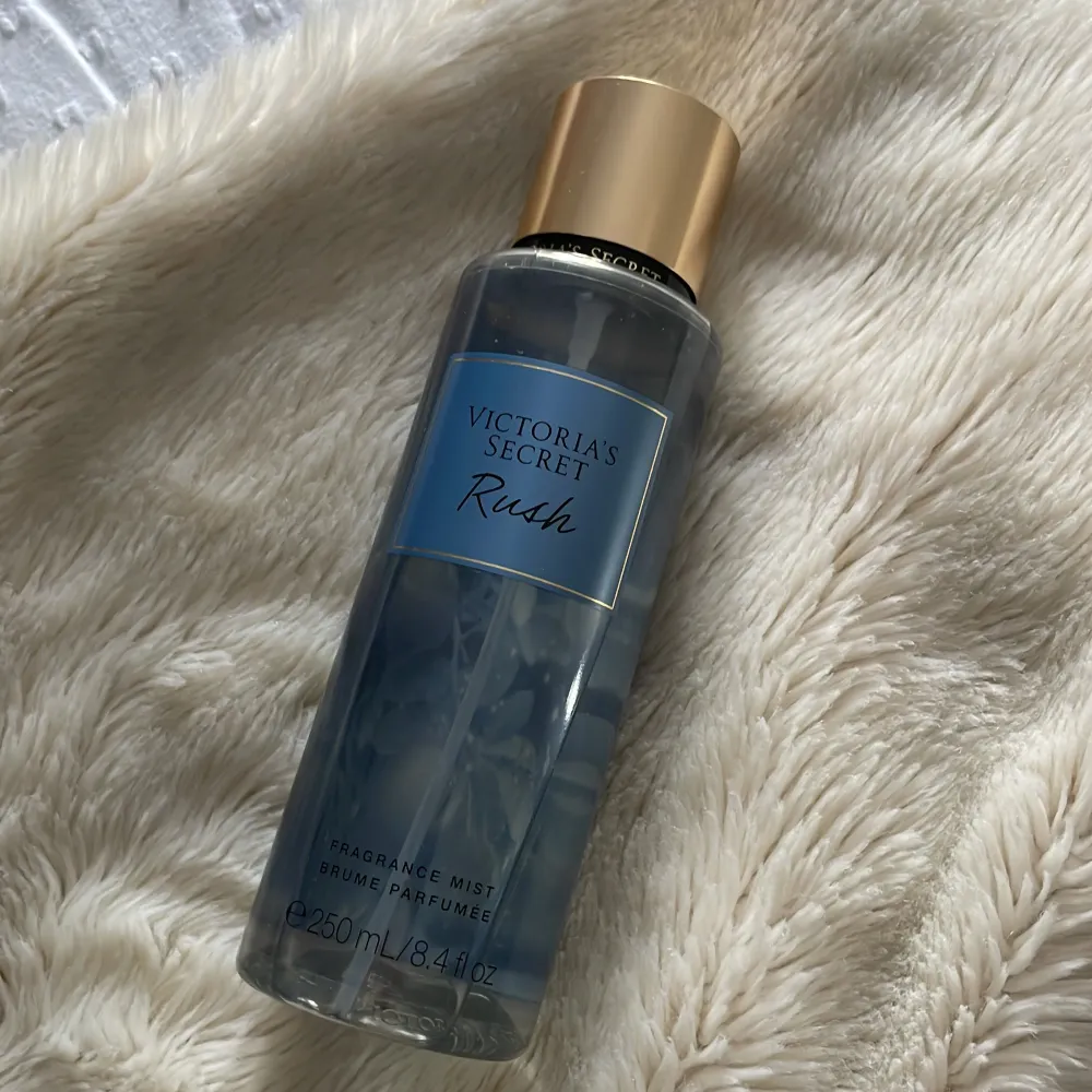 Säljer en Victoria's Secret Rush Fragrance Mist i en elegant genomskinlig flaska med blå etikett och guldfärgat lock. Innehåller 250 ml av en fräsch och uppfriskande doft. Perfekt för att ge en extra touch av lyx till din dagliga rutin.. Perfume.