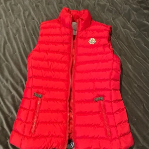 Röd dunväst från Moncler - Snygg röd dunväst från Moncler med dragkedja och två fickor framtill.Västen är i storlek S och passar även xs.