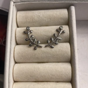 Silverfärgade örhängen med bladdetaljer - Eleganta silverfärgade örhängen formade som grenar med små blad och glittriga stenar. Perfekta för att ge en naturlig och stilren touch till din look.