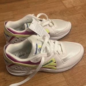 Vita sneakers från Reebok - Snygga vita sneakers från Reebok med färgglada detaljer i lila och gul. Skorna har en klassisk design med snörning och en bekväm sula. Perfekta för en sportig look. Och storleken är inte 36 