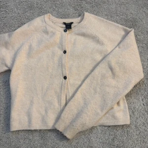 Beige kofta från Lindex  - Mysig beige kofta från Lindex med knappar framtill. Perfekt för kyligare dagar med sin mjuka och bekväma design. Långärmad och i en neutral färg som passar till det mesta. Ett måste i garderoben för en stilren look.