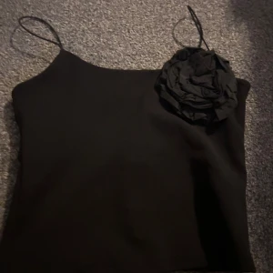 Svart axelbandslös topp med blomdetalj - Snygg svart axelbandslös topp med tunna axelband och en stor blomdetalj på ena sidan. Perfekt för en stilren och elegant look.