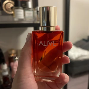Alive parfym intense  - En elegant parfymflaska med en djup röd färg och en stilren design. Flaskan har en gyllene kork och texten 'ALIVE' tryckt på framsidan. Perfekt för den som vill ha en sofistikerad doft.