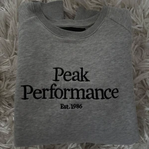 Grå sweatshirt från Peak Performance - Säljer en stilren grå sweatshirt från Peak Performance med svart text på bröstet. Tröjan har långa ärmar och ribbade muddar. Helt oanvänd